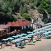 Un tratto di spiaggia con ombrelloni a righe e un ristorante sul lato, vicino a una scogliera verde.